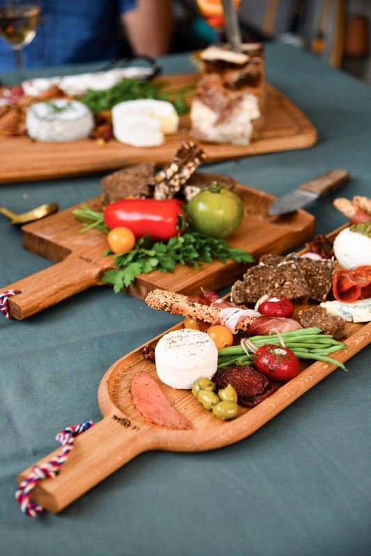 A LE RÉGAL - Lange Schaufel – großes Aperitifbrett aus Eichenholz mit Käse, Crackern, Brot, Kräutern, Oliven und Gemüse steht auf einem grünen Tischtuch. Im Hintergrund sind ein weiteres Brett, eine Person und eine nachhaltige Produktion zu erkennen.