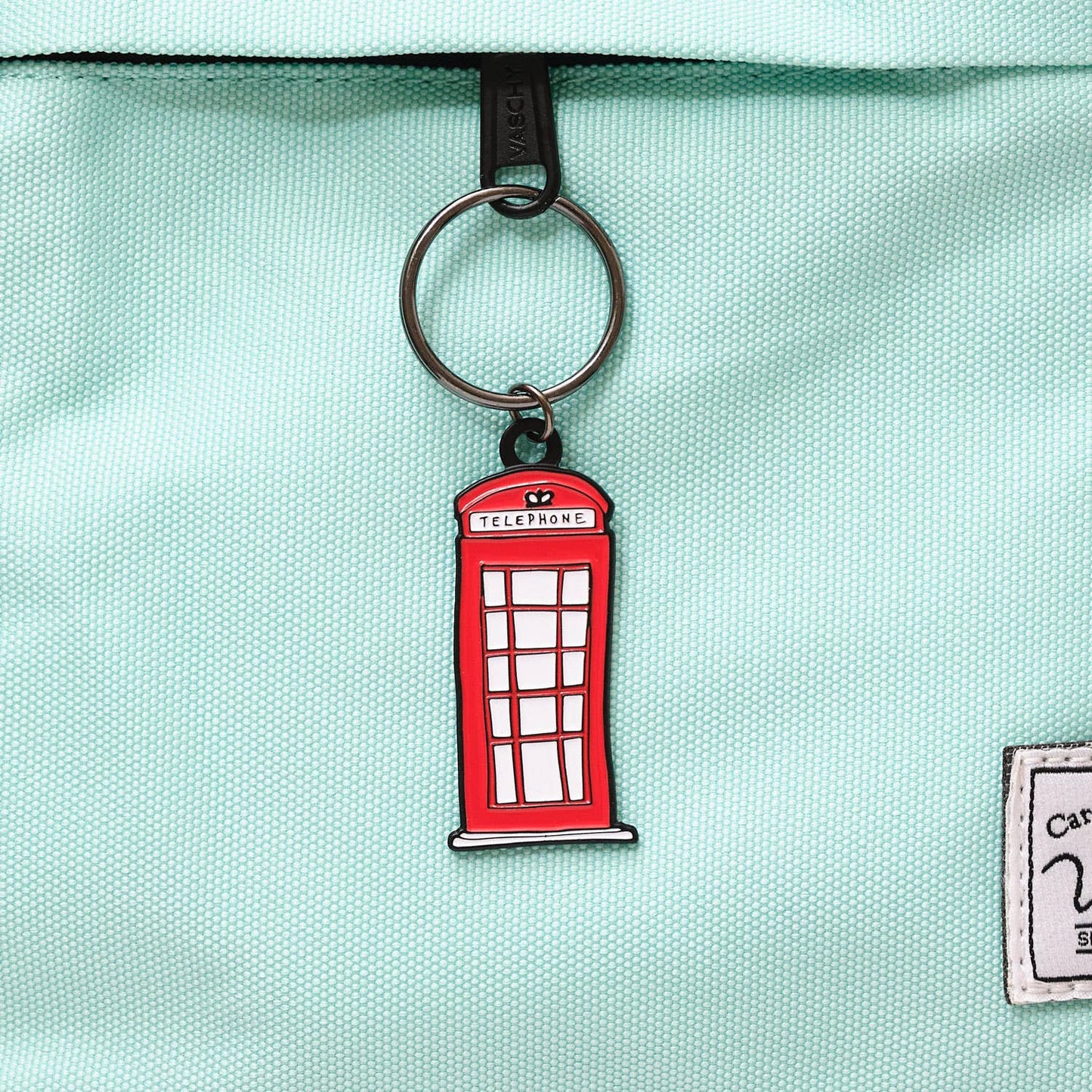 Der Victoria Eggs - Telefonzelle Schlüsselanhänger von Victoria Eggs ist am Reißverschluss einer hellblauen Stofftasche befestigt – das ideale Geschenk für London-Fans und Liebhaber britischer Designikonen.