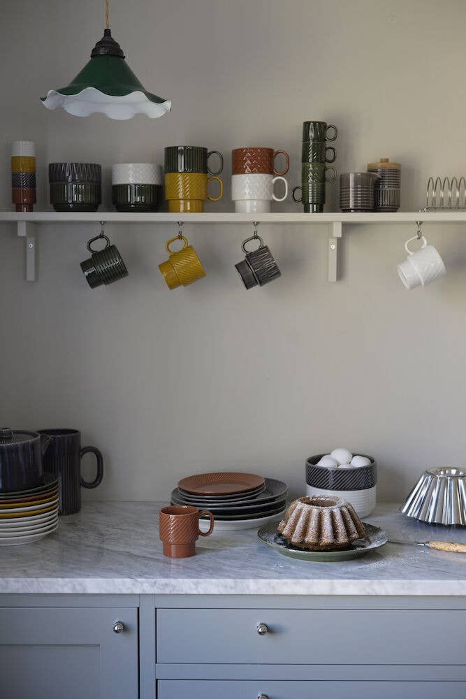 Eine Küche mit Marmorarbeitsplatten, blaugrauen Schränken, Tellern, einem Gugelhupf, Eiern und zweiteiligen Kaffeebechern der Sagaform GmbH Coffee & More – mit erdigen Tassen in den Regalen. Von der Decke hängt eine grüne Pendelleuchte.