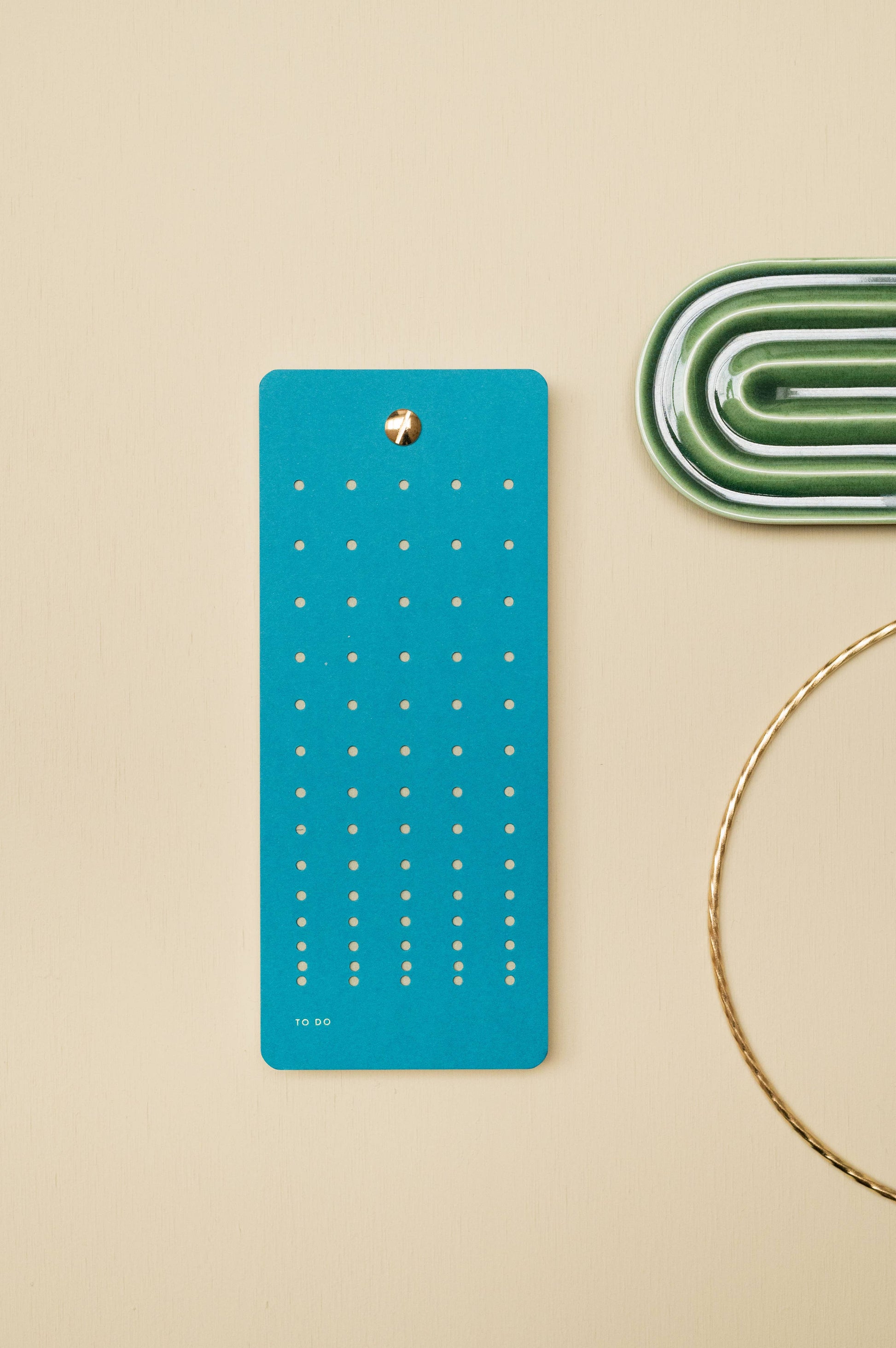 An einer beigen Wand hängt das blaugrüne Aufgabenlistenbuch "Tinne Mia - Aufgabenliste - Blaugrün" von Tinne Mia aus FSC®-Papier mit gepunkteten Reihen. Daneben stehen ein grünes Keramiktablett und ein goldener Drahtkreis auf cremefarbenem Hintergrund.