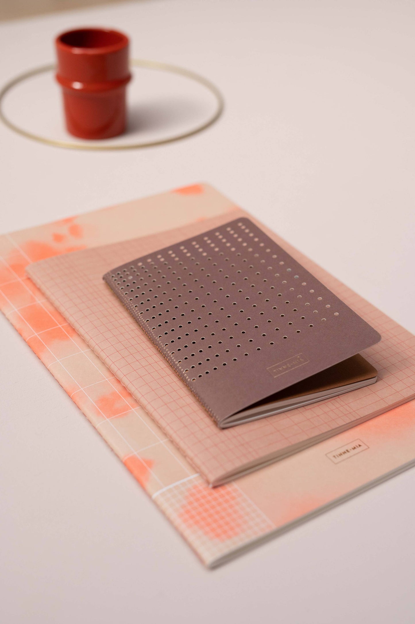 Drei Tinne Mia Bullet Journal A6 Notizbücher mit Rasterseiten und pastellfarbenen Einbänden sind auf einer weißen Fläche gestapelt. Das oberste Notizbuch ist die FSC Mix Edition "Burgunderrot". Im Hintergrund ruht eine rote Tasse auf einer weißen Untertasse.
