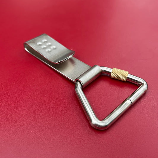 Der Niwaki-Gürtelclip ist ein dreieckiger Schlüsselanhänger aus Metall mit Karabinerhaken, einem strukturierten goldenen Schraubverschluss und einem flachen Clip, ideal zum Anbringen an einem Niwaki-Einzelholster oder als praktische Halterung, abgebildet auf einer leuchtend roten Oberfläche.