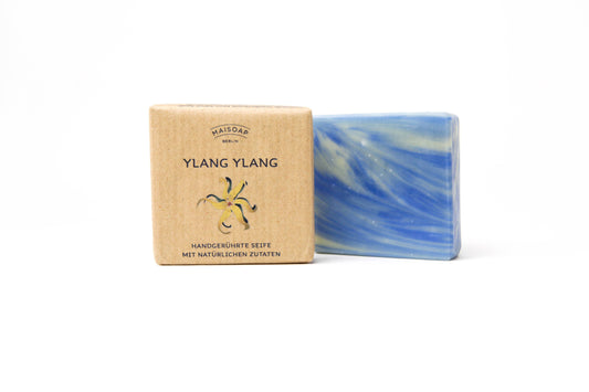 Die maisoap berlin - Ylang Ylang Seife liegt neben ihrer braunen, blumig gestalteten Verpackung auf weißem Hintergrund. Ihr weiß-blaues Marmorbild und blumig-zitroniger Duft sorgen für sanfte Hautpflege und ein besonderes Dufterlebnis.