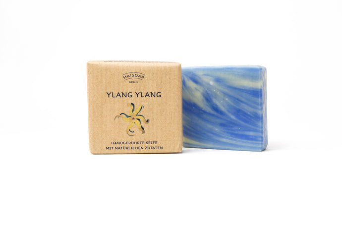 Die maisoap berlin - Ylang Ylang Seife liegt neben ihrer braunen, blumig gestalteten Verpackung auf weißem Hintergrund. Ihr weiß-blaues Marmorbild und blumig-zitroniger Duft sorgen für sanfte Hautpflege und ein besonderes Dufterlebnis.