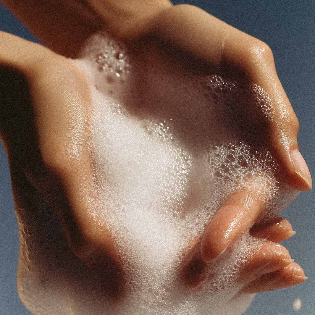 Zwei Hände, bedeckt mit weißen, schaumigen Seifenblasen vor einem zartblauen Hintergrund, suggerieren eine Handwäsche mit Panier Des Sens Duftseife | Zeste Riviera 150 g.