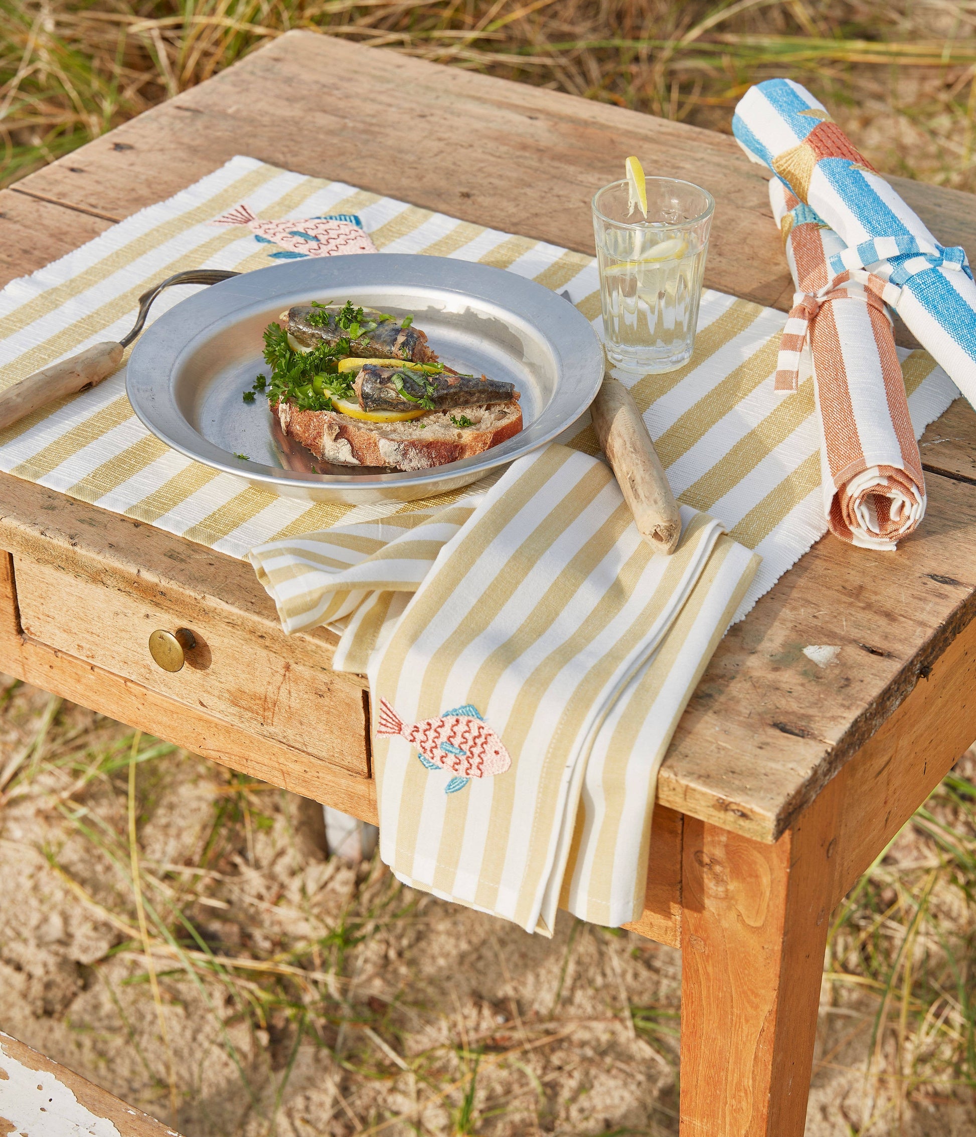 Auf einem Holztisch im Freien stehen eine Serviette von Sylvie Thiriez, ein Teller Fisch auf Brot mit Kräutern, Besteck und ein Glas Zitronenwasser. Zwei gerollte gestreifte Servietten von Sylvie Thiriez liegen ebenfalls bereit. Im Hintergrund sind Gras und Sand zu sehen.