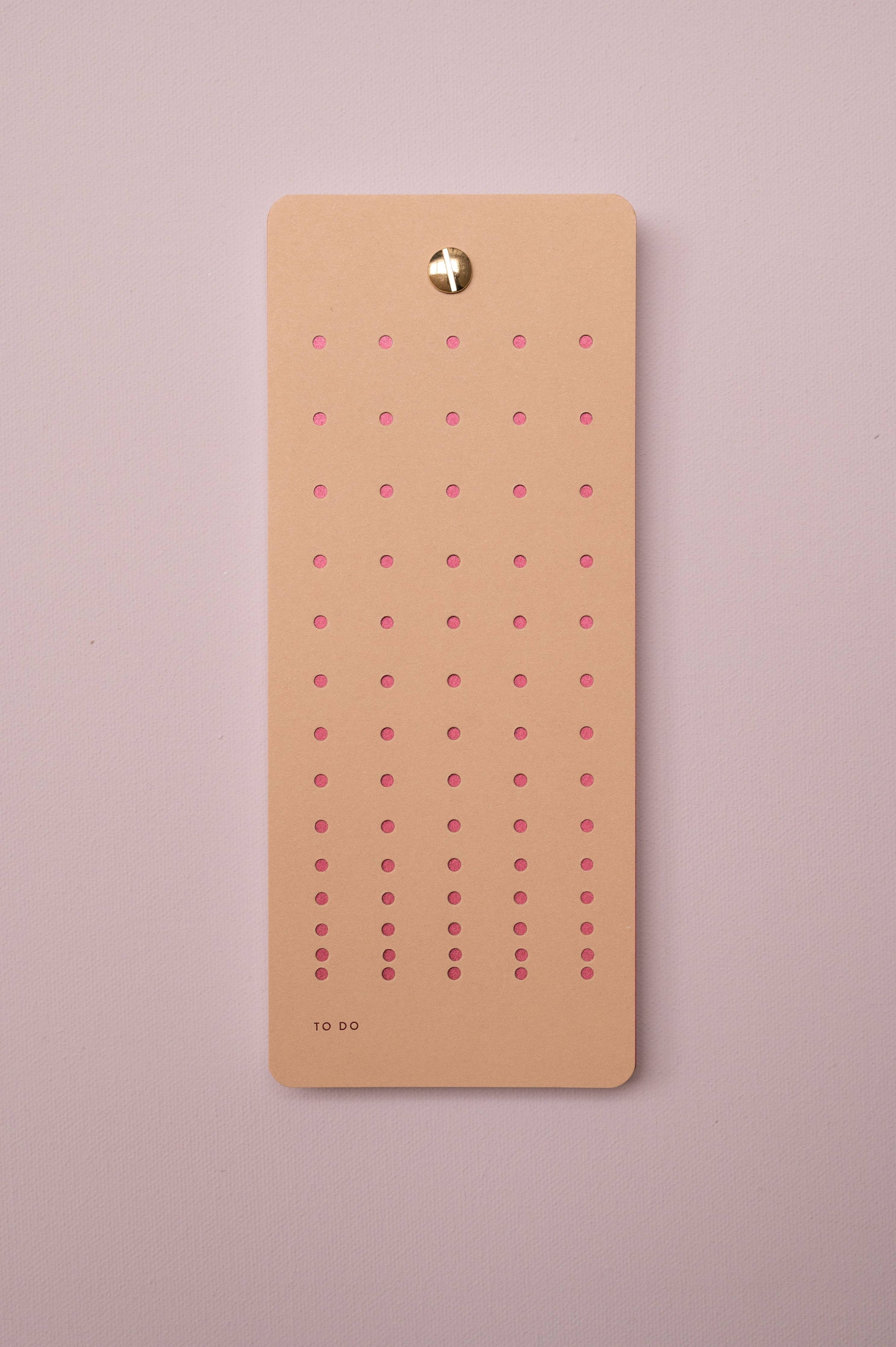 Die Tinne Mia Aufgabenliste "Nougat" aus FSC-zertifiziertem Papier mit sechs rosafarben gepunkteten Spalten ist an einer zartrosa Wand befestigt. Unten links auf der Karte steht klein "TO DO".