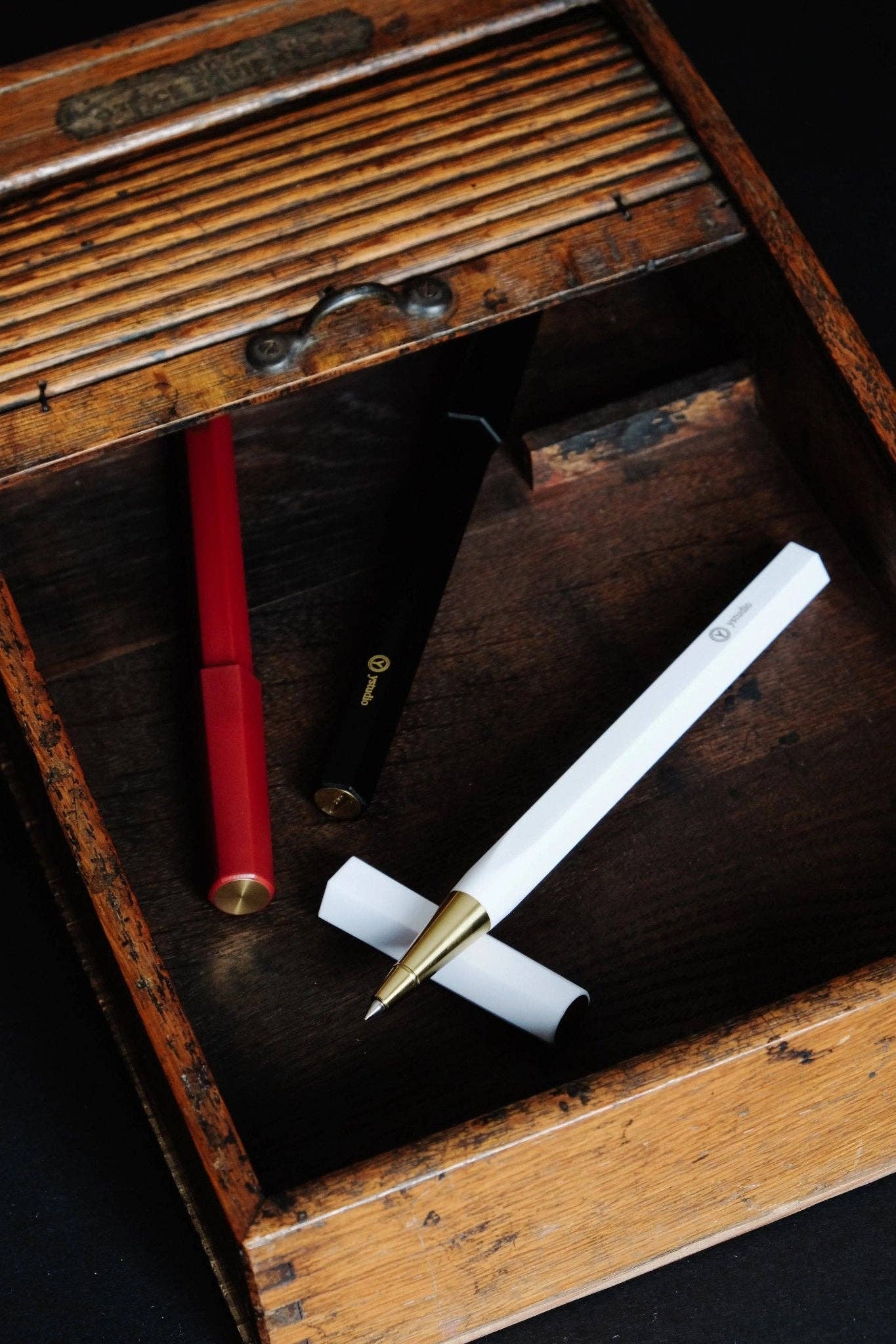 Der ENTREPÔT - YSTUDIO Harz-Rollerball-Stift (Schwarz) von ENTREPÔT liegt zusammen mit roten und weißen Stiften in einer rustikalen Holzschublade mit Lattenabdeckung vor einem dunklen Hintergrund.