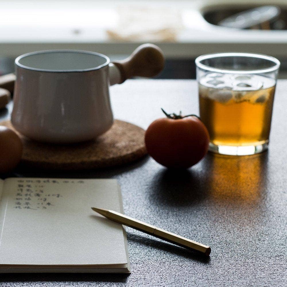 Ein ENTREPÔT - YSTUDIO - Klassischer Revolve Kugelschreiber Schlank ruht auf einem aufgeschlagenen Notizbuch mit handschriftlichen Notizen, neben einer Tomate, einem Glas Eistee und einer weißen Kanne auf einer Korkmatte, alles auf einer dunklen Arbeitsplatte.