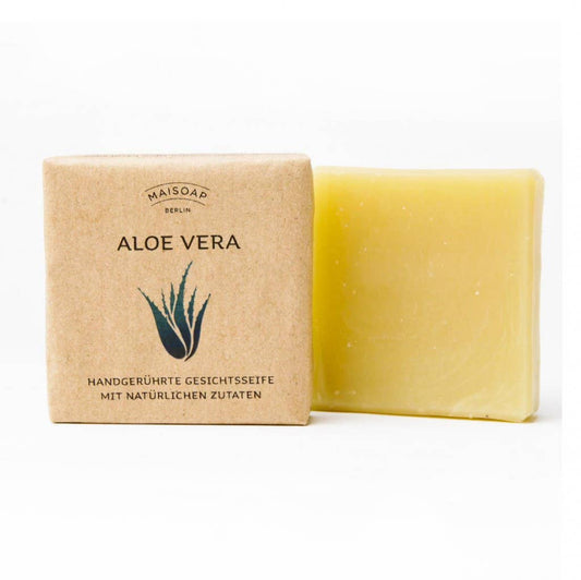 Ein beiger Riegel maisoap berlin - Aloe Vera Seife ruht neben seiner braunen Papierverpackung, bedruckt mit einer grünen Aloe-Pflanze und einem deutschen Text, der die natürlichen Inhaltsstoffe und die handwerkliche Herstellung für eine sanfte Gesichtshaut betont.