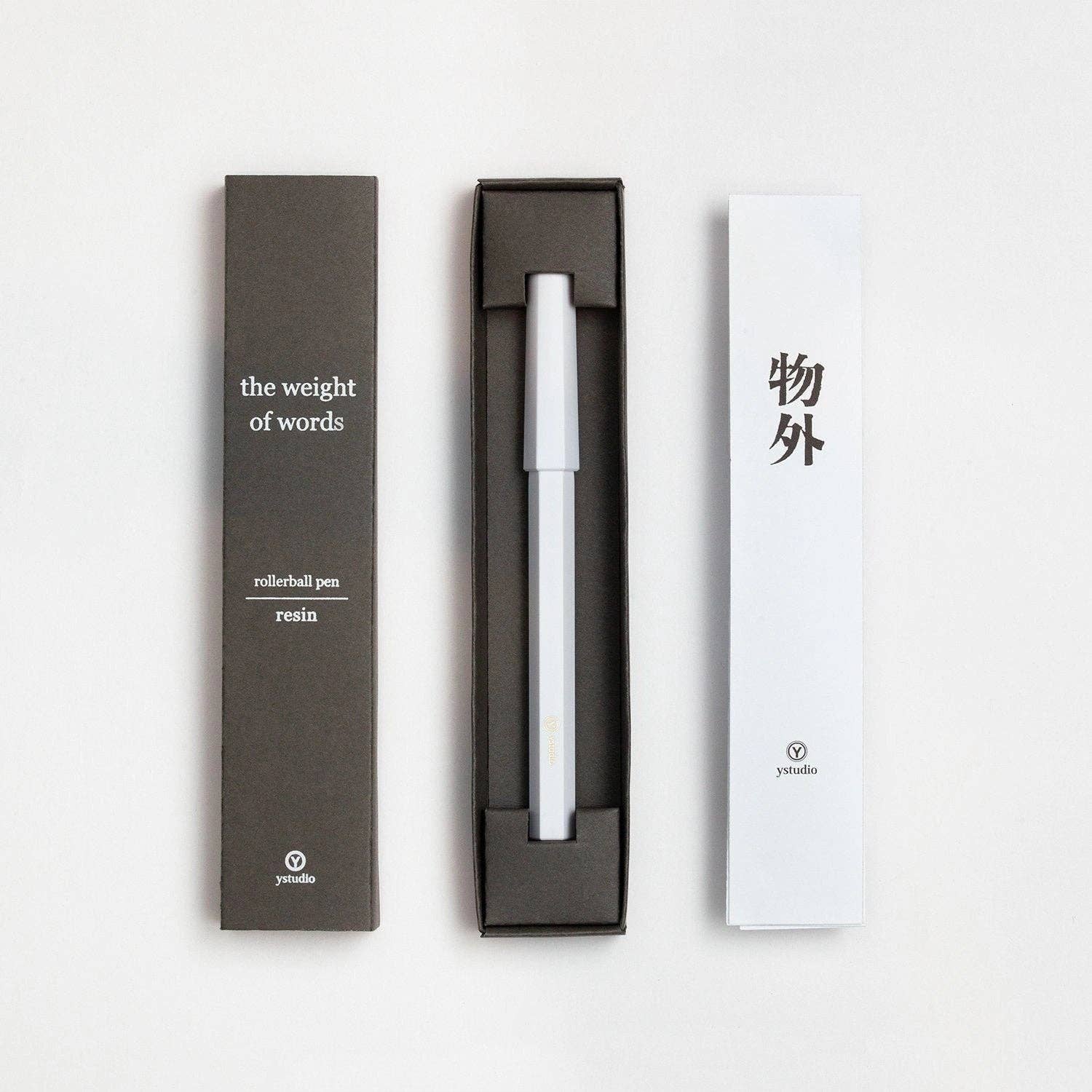 Der ENTREPÔT - YSTUDIO Harz-Rollerball-Stift (Schwarz) von ENTREPÔT wird in einer schwarzen Schachtel gezeigt, platziert zwischen seiner Verpackung "the weight of words, rollerball pen, Acryl und Messing" und einer weißen Karte mit asiatischen Schriftzeichen und Logo.