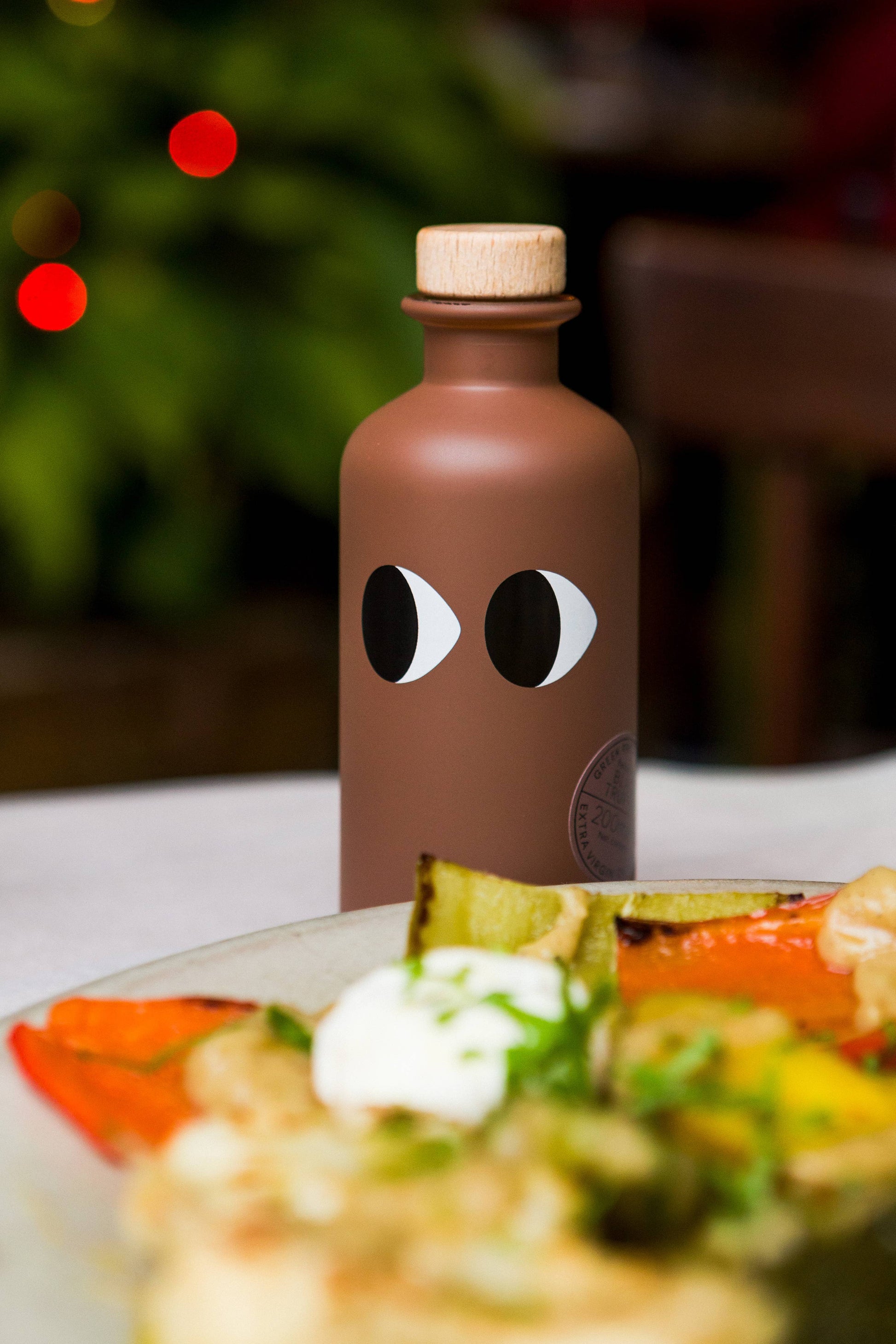 Eine braune "yiayia and friends" Natives Olivenöl extra mit Trüffel Flasche mit Cartoon-Augen steht auf einem Tisch, neben einem Teller mit buntem gegrilltem Gemüse und Sahne, mit verschwommenen Lichtern und Grünzeug im Hintergrund.