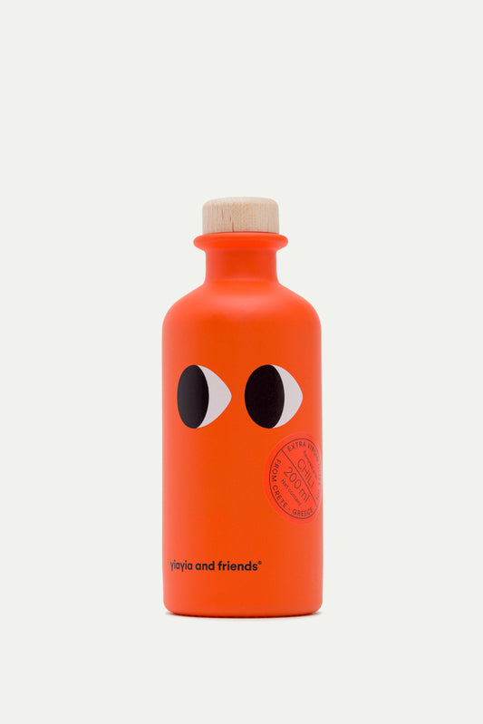 In der Nähe des Bodens befindet sich eine leuchtend orangefarbene Flasche mit einem Holzdeckel, zwei Cartoon-Augen und einem Stempel mit der Aufschrift "Natives Olivenöl extra mit Chili" von yiayia and friends.