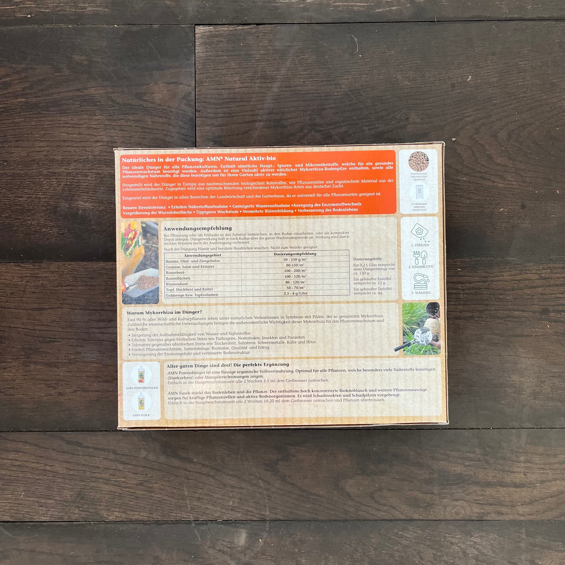 Auf der Rückseite einer AMN Natural Aktiv-bio-Verpackung finden sich deutscher Text, Nährwertangaben und Anwendungshinweise. Auf dunklem Holzboden zeigen kleine Bilder Pflanzen und Gartenarbeit und weisen die Packung als AMNs Gesunddünger mit Mykorrhiza aus.