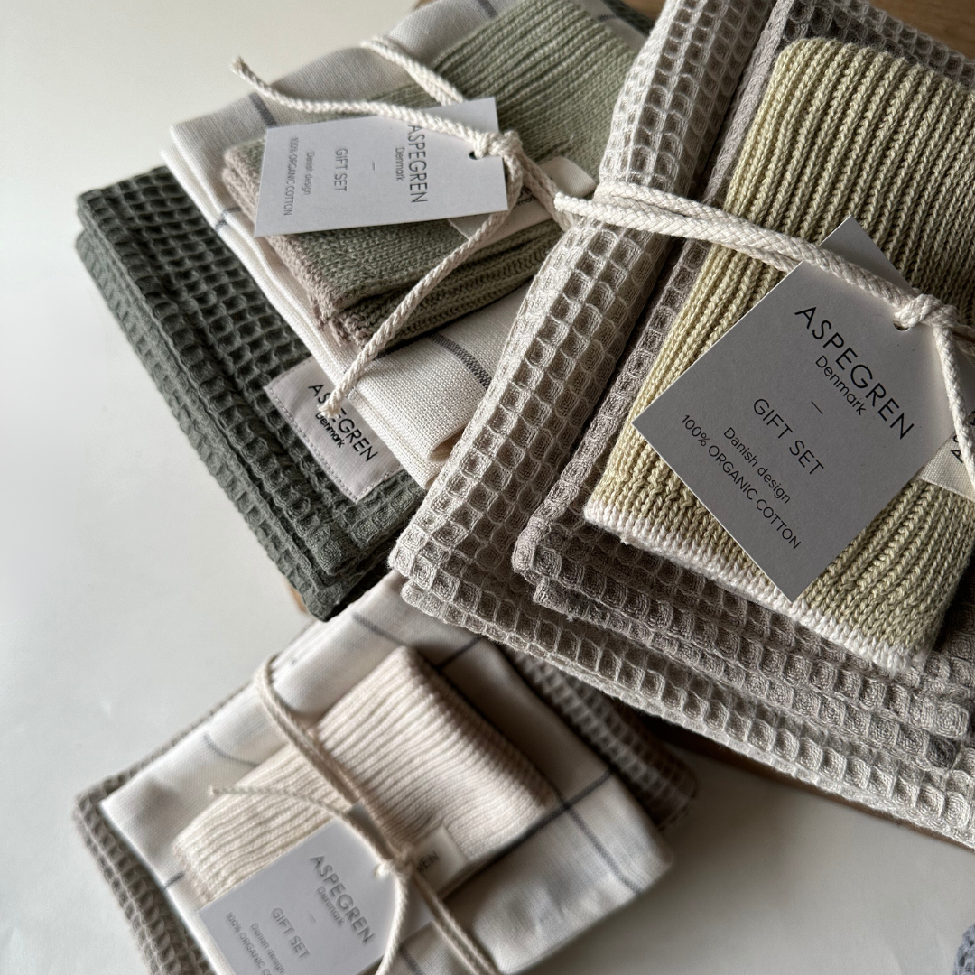 Mehrere ordentlich gefaltete Handtücher „Aspegren Denmark Geschenkset – Lemon Grass“ in Grün-, Beige- und Cremetönen aus 100 % Bio-Baumwolle sind mit Etiketten auf einer weißen Oberfläche gebündelt.