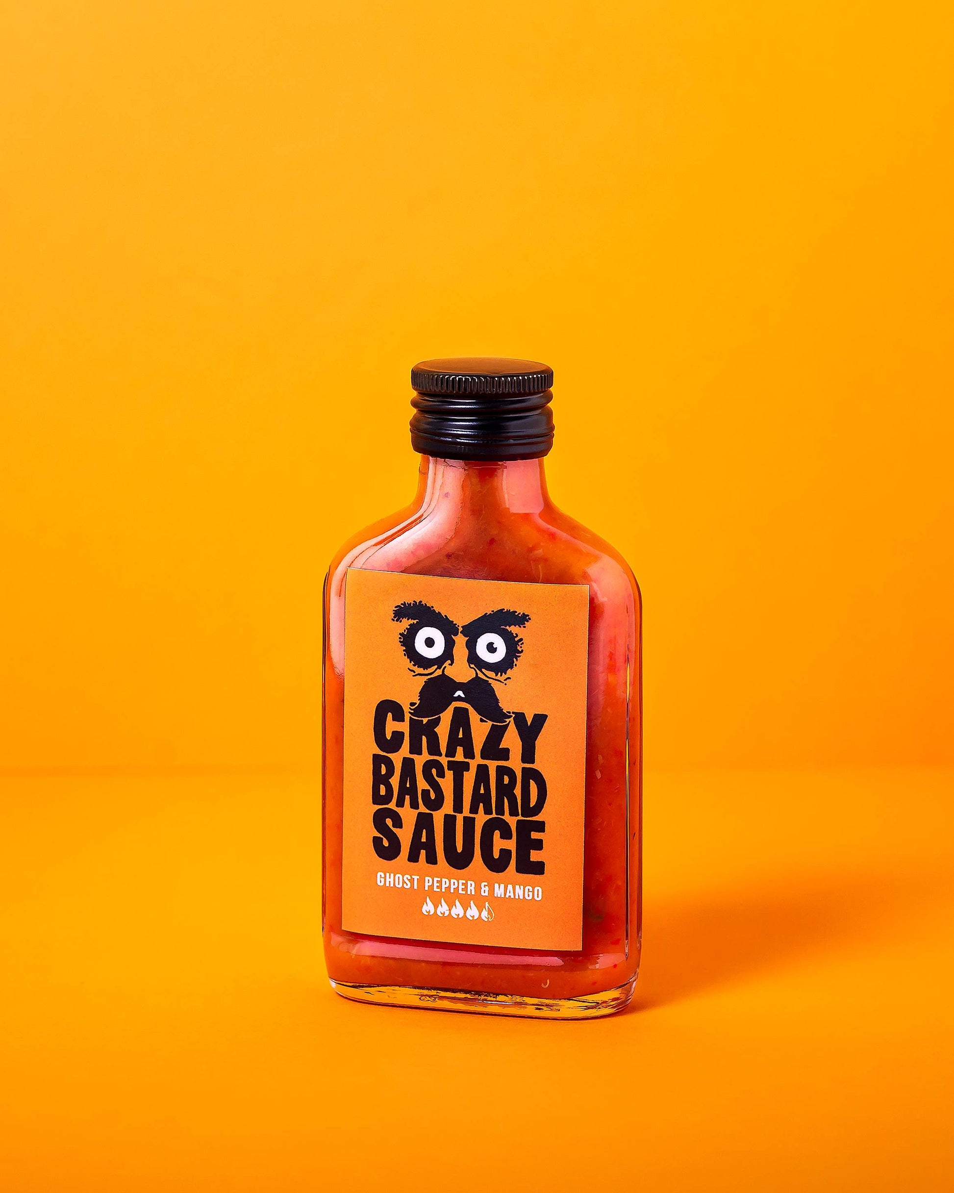 Die rechteckige Glasflasche von CBS Foods GmbH (Crazy Bastard Sauce) - Ghost Pepper & Mango 100 ml scharfe Soße hat ein orangefarbenes Etikett mit fetter schwarzer Schrift und Cartoon-Augen vor einem leuchtend orangen Hintergrund.