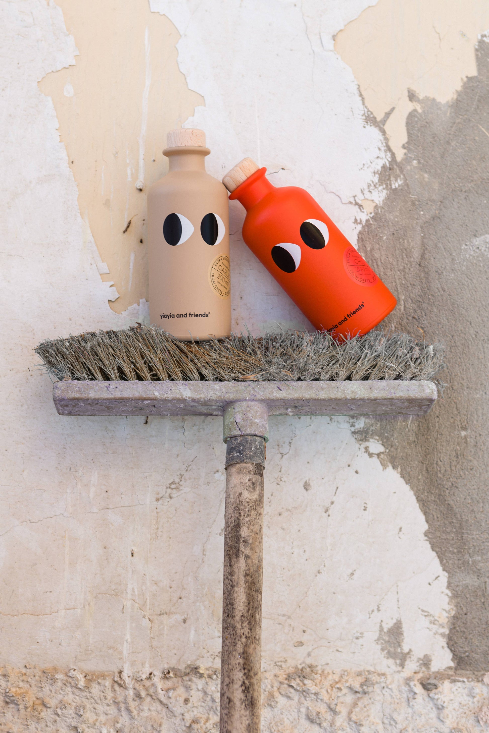 Zwei Flaschen von yiayia and friends - eine beige, eine orangefarbene mit minimalistischen Gesichtern - stehen auf alten Besenstielen an einer abblätternden Wand; eine enthält das yiayia and friends Natives Olivenöl extra mit Chili.