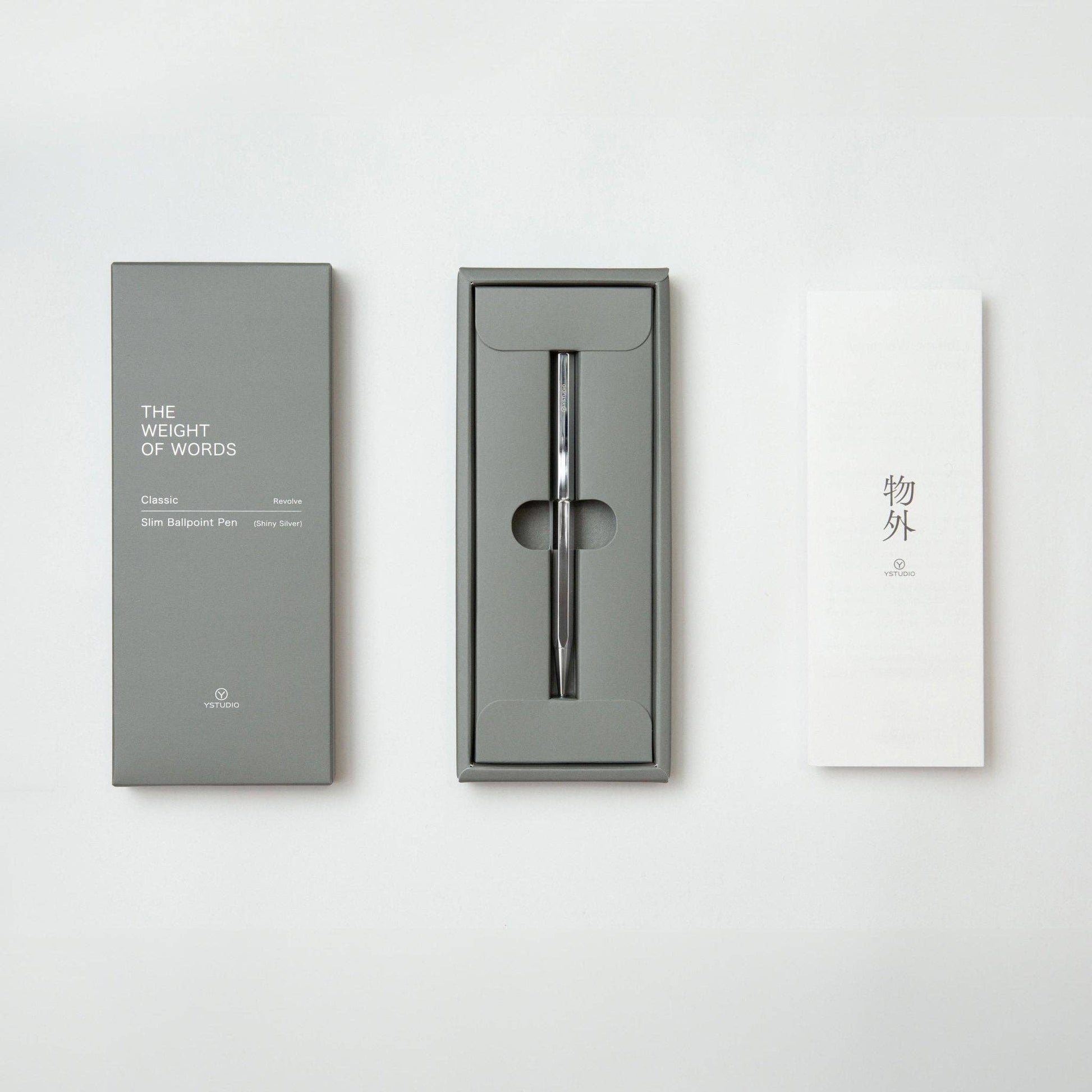 Der ENTREPÔT - YSTUDIO Klassischer Revolve Kugelschreiber Slim (Glänzendes Silber) liegt in einer grauen Box zwischen der Verpackung "The Weight of Words" links und einem weißen Papier mit asiatischen Zeichen rechts.
