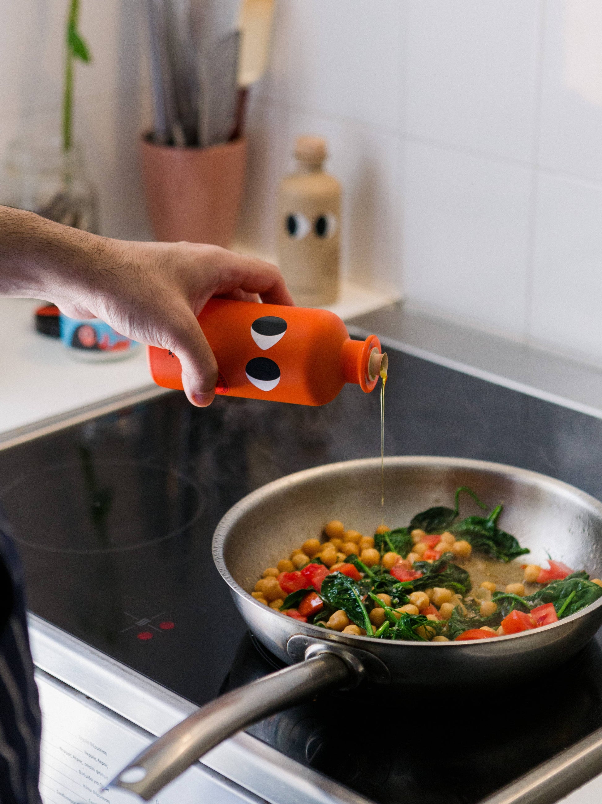 Eine Hand gießt yiayia and friends - Natives Olivenöl extra mit Chili aus einer orangefarbenen Flasche mit schwarzen und weißen Augen auf Kichererbsen, Spinat, rote Paprika und Chilischoten, die in einer Pfanne auf dem Herd brutzeln.