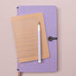 Ein Tinne Mia Notizbuch A6 - Nougat Grid (FSC Mix) und ein weißer Stift befinden sich auf einem geschlossenen lavendelfarbenen Journal mit Knopfverschluss vor einem hellrosa Hintergrund.