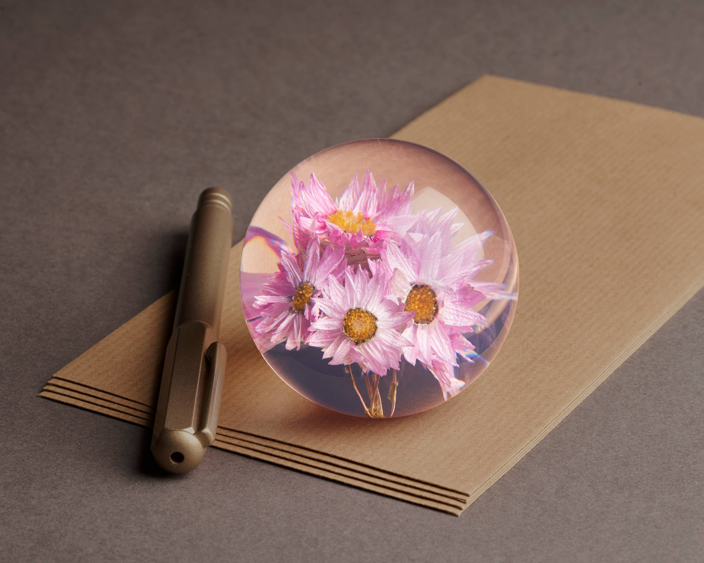 Ein Metallstift und braune Umschläge liegen auf einem Tisch, auf dem ein Hafod Grange Rhodanthe Briefbeschwerer liegt - ein dekorativer Briefbeschwerer mit rosa und gelben Blüten.