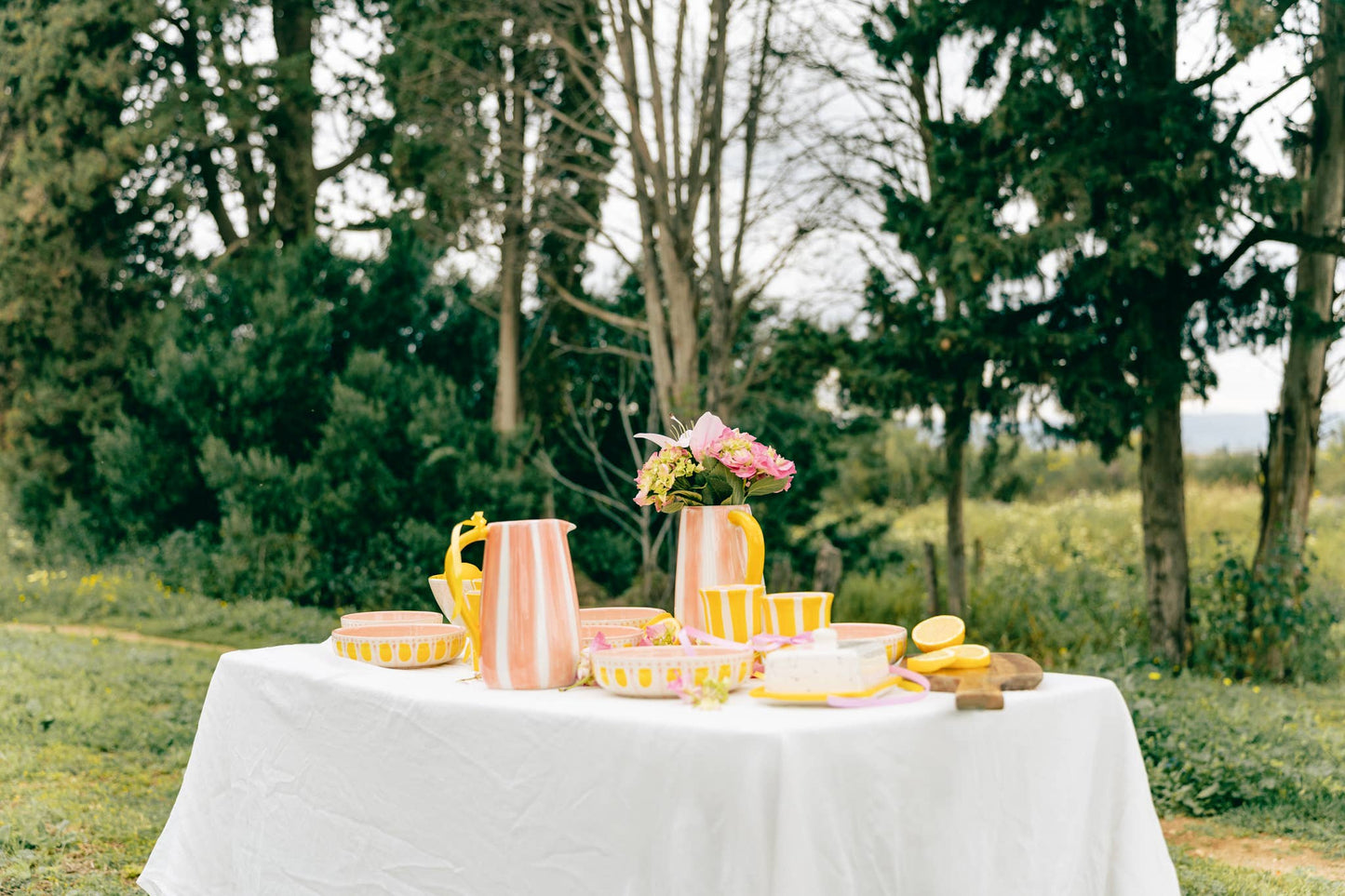 Ein weiß gedeckter Tisch im Freien mit der handgefertigten Keramik-Servierschale 1,5L Rosa und Gelb VALENTINA von Alfar Tierra Cocida, rosa und gelbem Geschirr, einer Vase mit Blumen, Zitronen und Bäumen im Hintergrund.