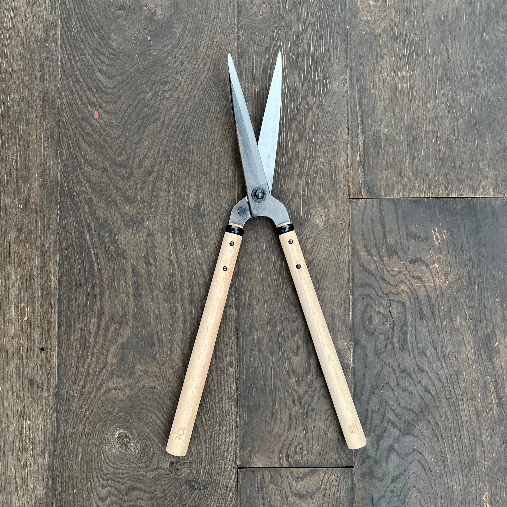 Ein Paar Niwaki FSC Heckenschere von Niwaki mit Holzgriffen aus FSC-zertifiziertem Holz und offenen Metallklingen ruht auf einem dunklen Holzboden.