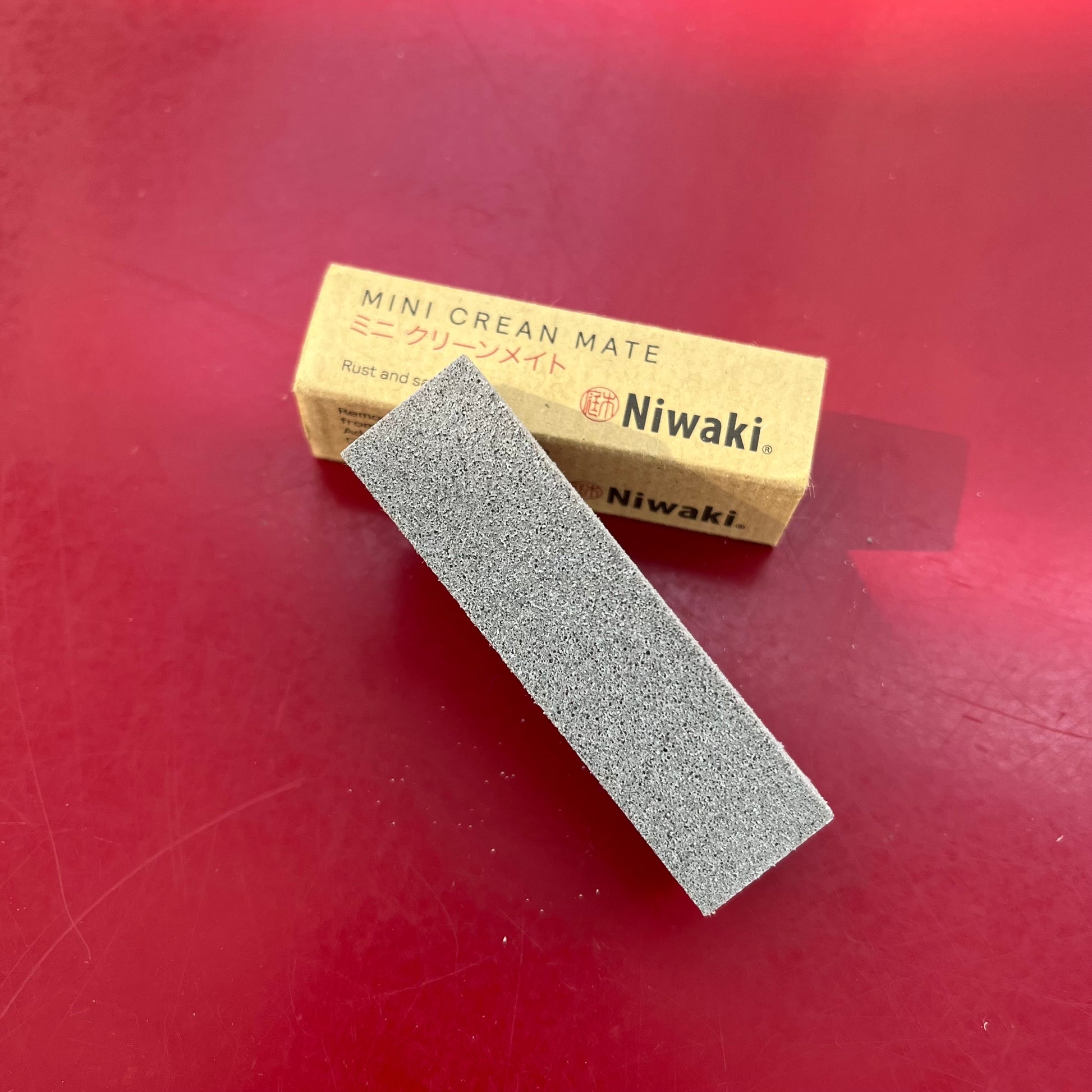 Ein grauer rechteckiger Schärfstein-Reinigungsblock sitzt auf einer kleinen braunen Niwaki - Mini Crean Mate Box, ideal zum Schärfen von Gartenscheren, ausgestellt auf einer roten Fläche.