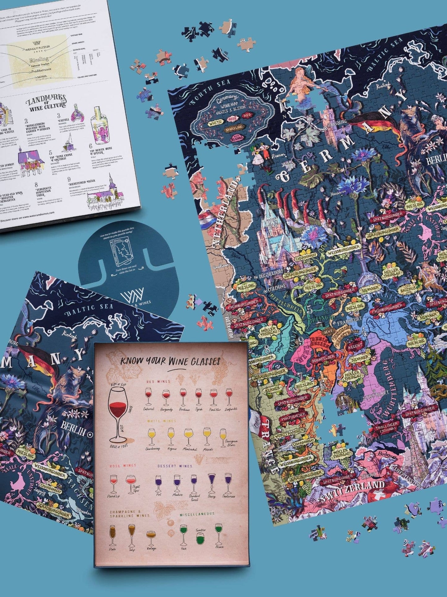 Ein teilweise ausgefülltes Water & Wines - Wein Puzzle - Deutschland liegt auf einer blauen Fläche mit losen Teilen in der Nähe, zusammen mit illustrierten Weinregionsführern und einer Karte mit Weingläsern, die deutsche Weinregionen hervorheben.