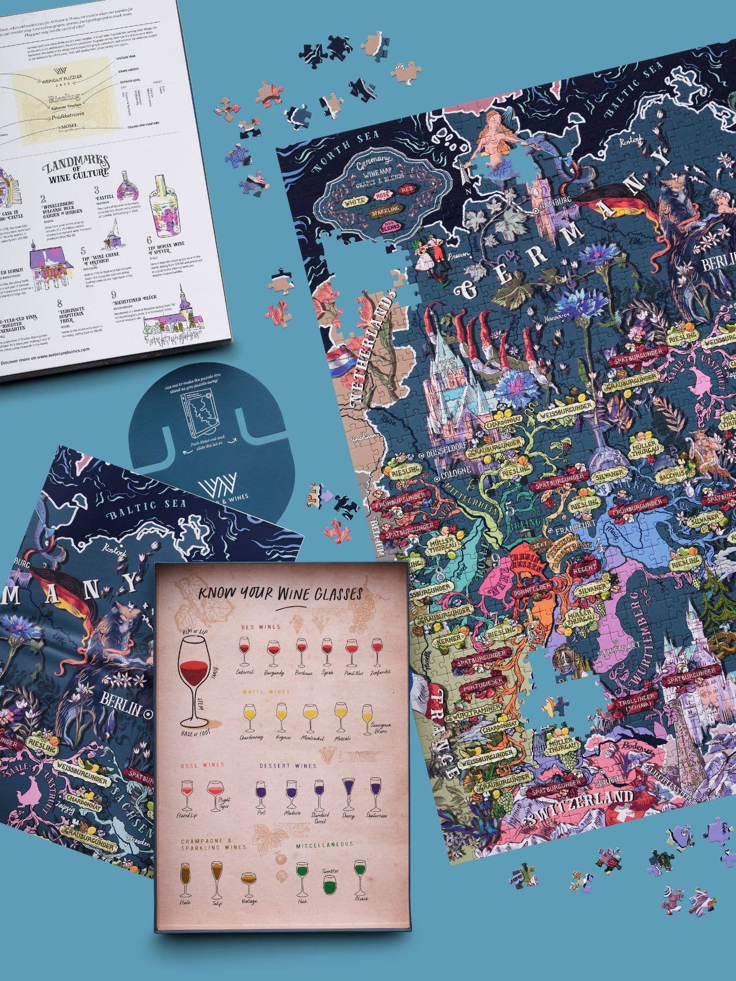 Ein teilweise ausgefülltes Water & Wines - Wein Puzzle - Deutschland liegt auf einer blauen Fläche mit losen Teilen in der Nähe, zusammen mit illustrierten Weinregionsführern und einer Karte mit Weingläsern, die deutsche Weinregionen hervorheben.