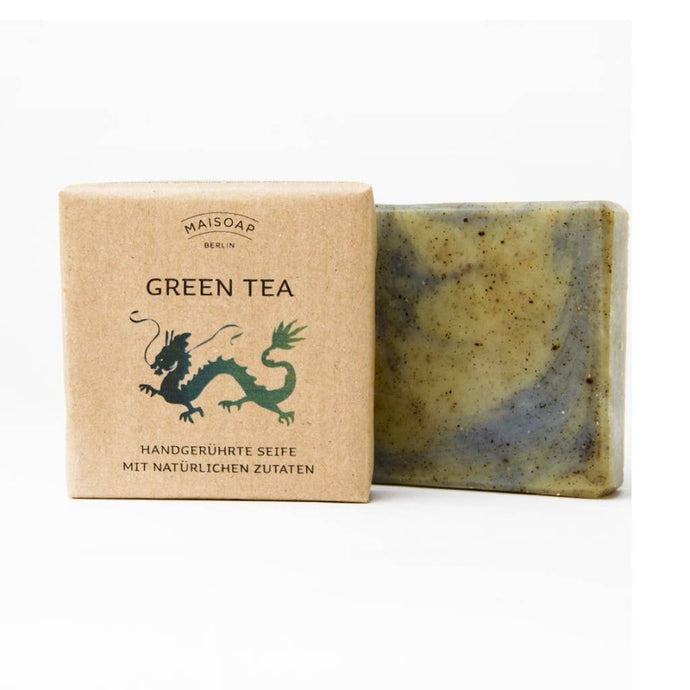 Ein Riegel maisoap berlin - Grüner Tee Seife mit sichtbaren Kräutern steht neben seiner braunen Papierverpackung, auf der ein grüner Drache und die Worte 