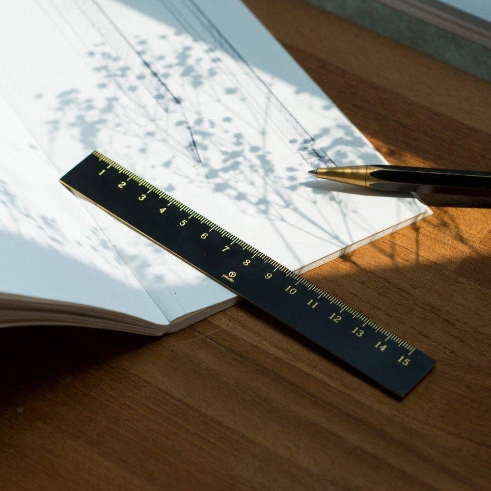 Ein ENTREPÔT YSTUDIO Klassischer Reflect Lineal, gepaart mit einem Stift und einem aufgeschlagenen Notizbuch im Schatten eines Baumes, elegant auf einer Holzfläche arrangiert.