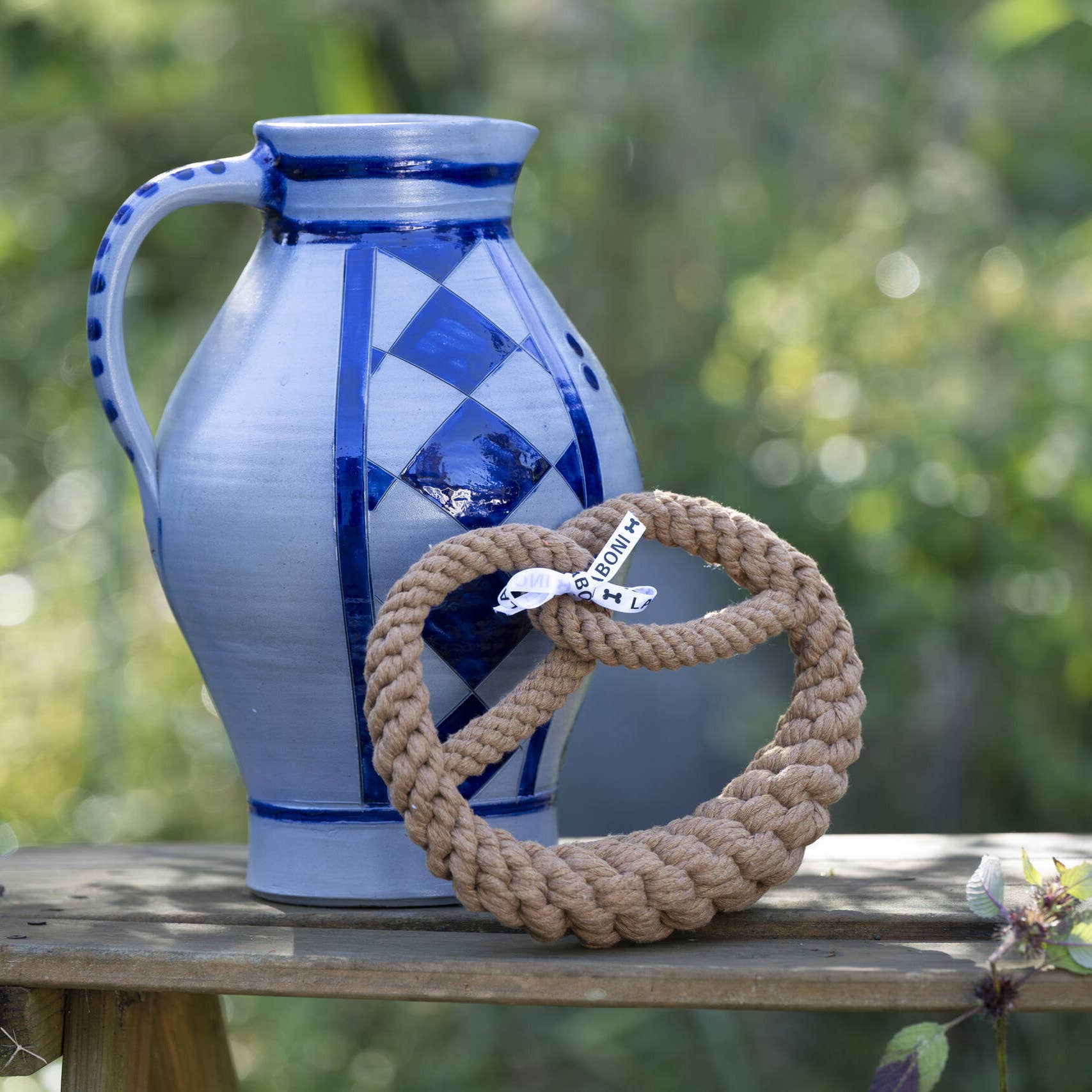 Eine blaue und graue Keramikkanne mit geometrischen Mustern steht auf einer hölzernen Oberfläche im Freien, neben dem Untersetzer LABONI - Volentis GmbH - Britta Brezel aus geflochtenem Baumwollseil, der ein kleines weißes Etikett trägt. Grünes Laub ist im Hintergrund verschwommen zu erkennen.