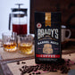 Ein Beutel Brady's Coffee Company - Brady's Barrel Aged Irish Whiskey Kaffeebeutel mit ganzen Bohnen ruht auf einem Holzbrett. Dahinter erinnern zwei Gläser, eine Kaffeemaschine und eine Tasse an die reiche Erfahrung des Whiskey-Kaffeegenusses.