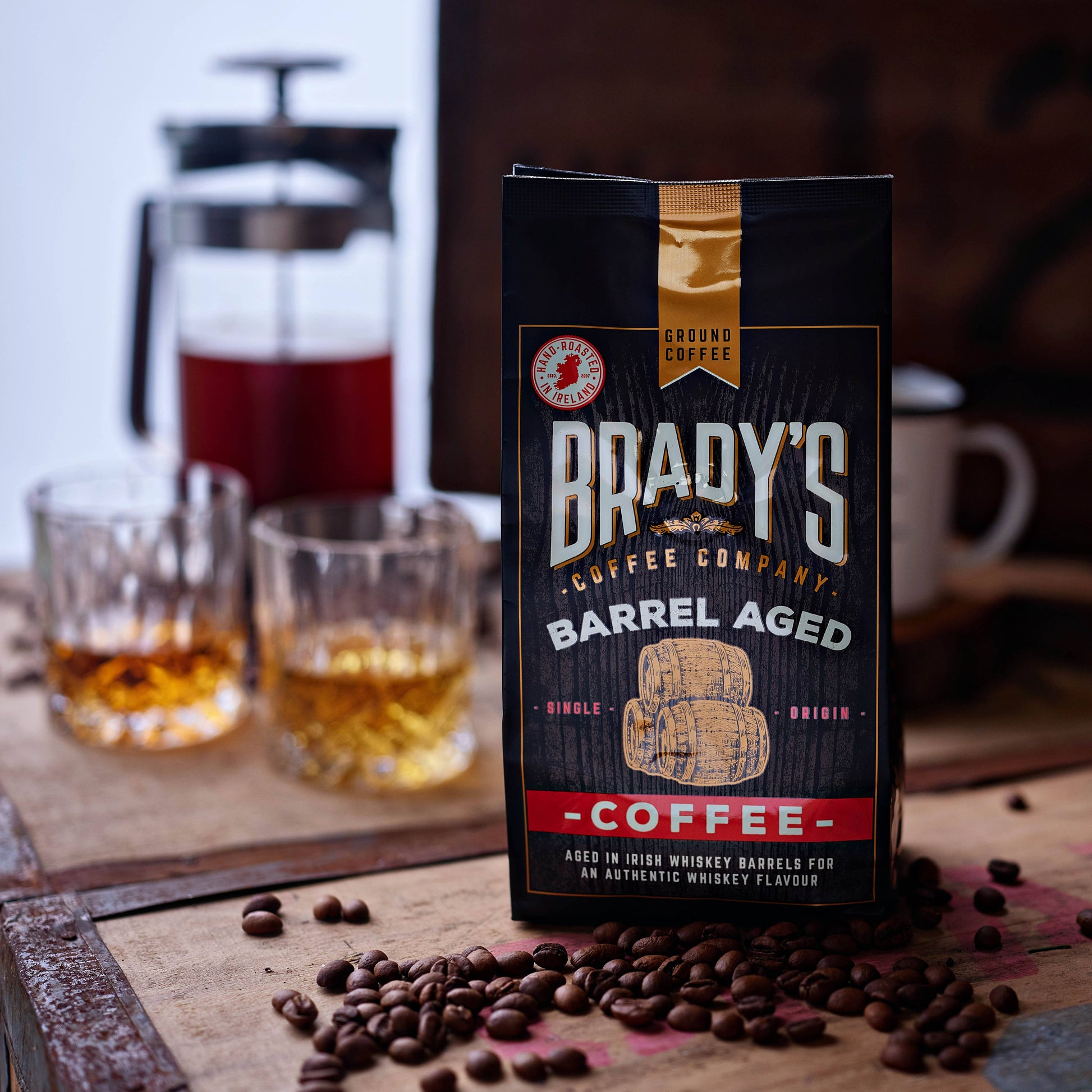 Ein Beutel Brady's Coffee Company - Brady's Barrel Aged Irish Whiskey Kaffeebeutel mit ganzen Bohnen ruht auf einem Holzbrett. Dahinter erinnern zwei Gläser, eine Kaffeemaschine und eine Tasse an die reiche Erfahrung des Whiskey-Kaffeegenusses.