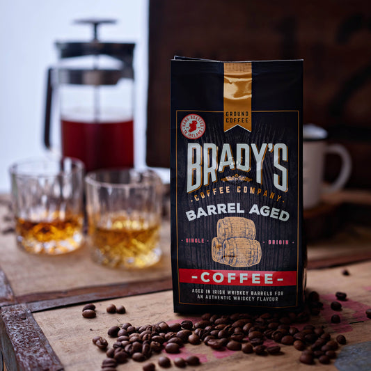 Ein Beutel Brady's Coffee Company - Brady's Barrel Aged Irish Whiskey Kaffeebeutel mit ganzen Bohnen ruht auf einem Holzbrett. Dahinter erinnern zwei Gläser, eine Kaffeemaschine und eine Tasse an die reiche Erfahrung des Whiskey-Kaffeegenusses.