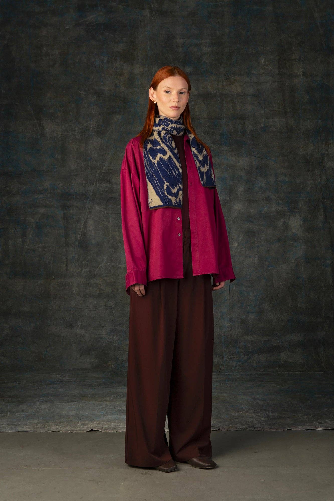 Eine Frau mit langen roten Haaren steht vor einem dunklen, strukturierten Hintergrund. Sie trägt ein lockeres magentafarbenes Hemd, weite braune Hosen, braune Schuhe und den Wolvis Schal aus Merinowolle - Aurata Indigo Mini leger über die Schultern gelegt.