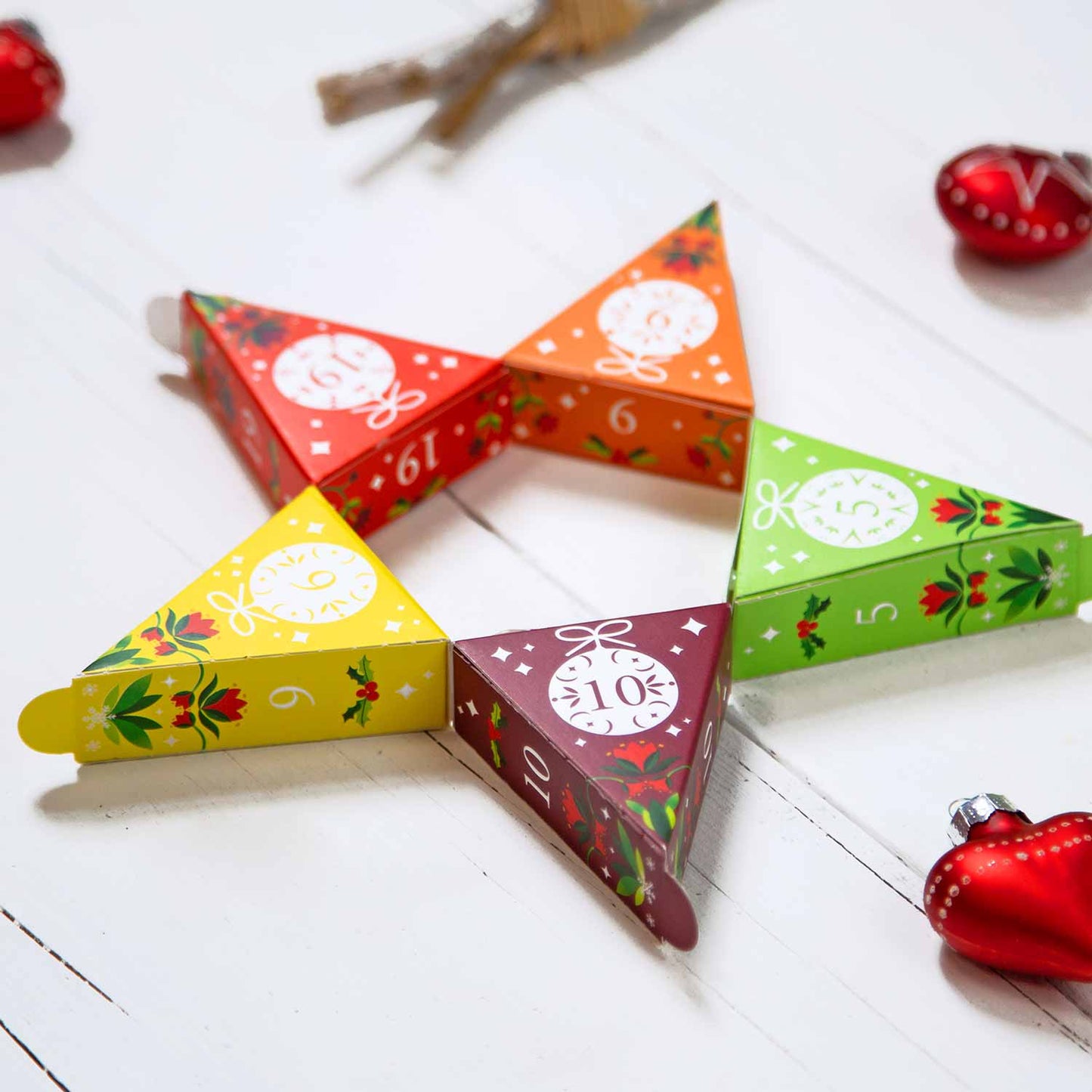 Fünf bunte, dreieckige Schachteln aus dem English Tea Shop BIO Tee Adventskalender "Christmas Vibes only" (25 Pyramidenbeutel) sind sternförmig auf einer weißen Holzfläche angeordnet, jede nummeriert und festlich verziert, mit roten Herz-Ornamenten in der Nähe.