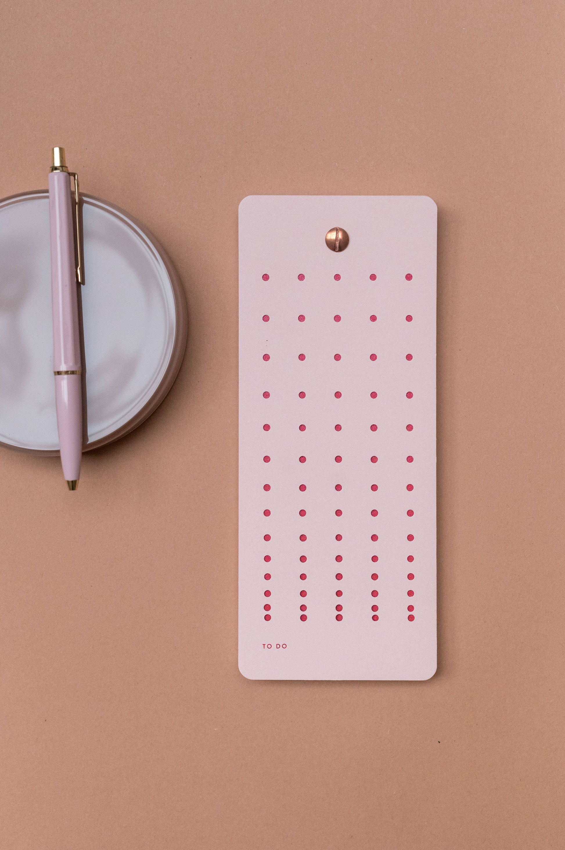 Die Aufgabenliste "Mellow Rose" von Tinne Mia aus FSC®-zertifiziertem Papier mit roten Punkten liegt neben einem rosa-goldenen Stift auf einer weißen Schale - alles nachhaltig produziert und stilvoll arrangiert.