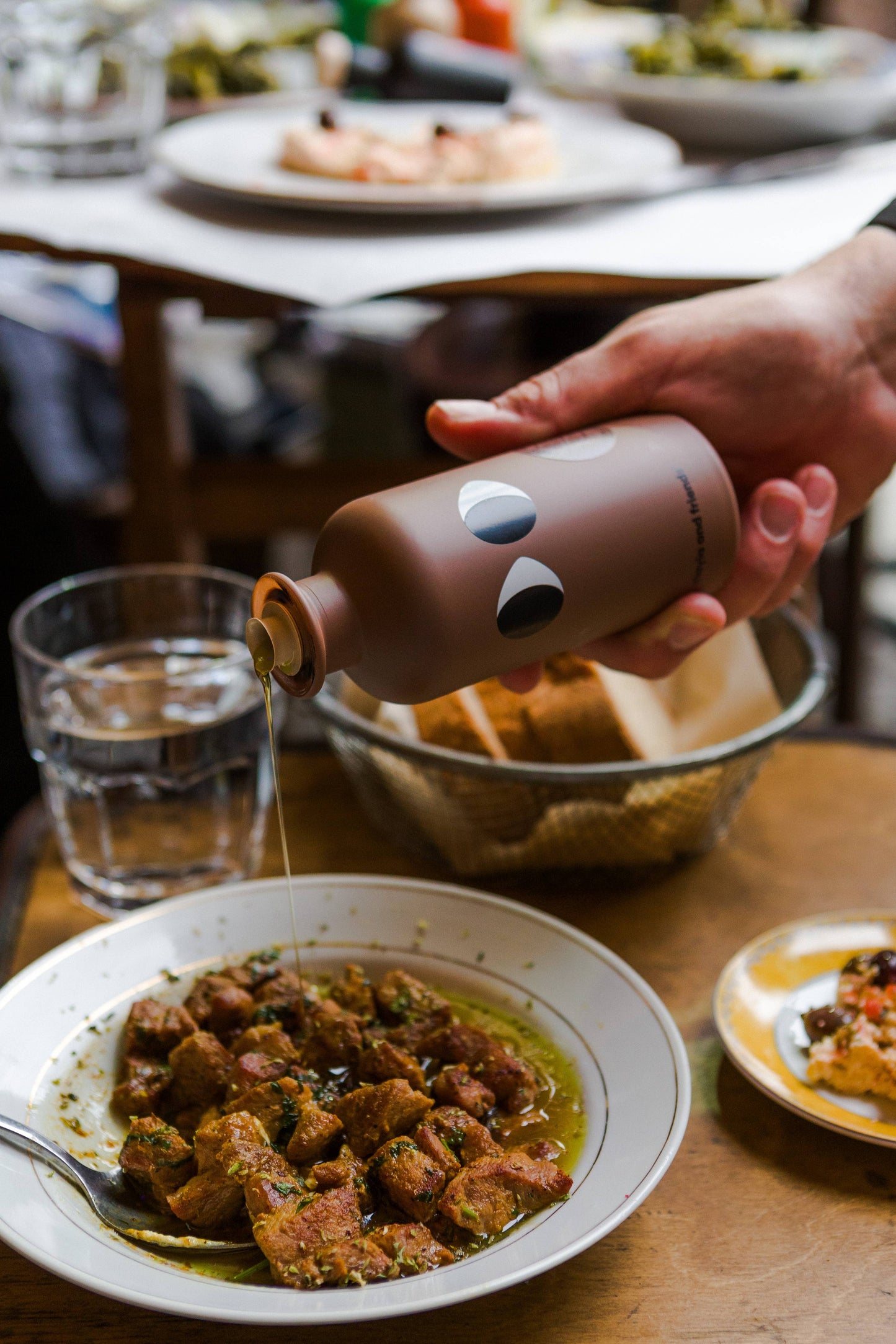Eine Hand schüttet yiayia and friends - Natives Olivenöl extra mit Trüffel aus einer stilvollen Flasche auf marinierte Fleischwürfel. Auf dem Tisch stehen ein Glas Wasser, Brot in einem Korb und Teller mit Essen im Hintergrund.