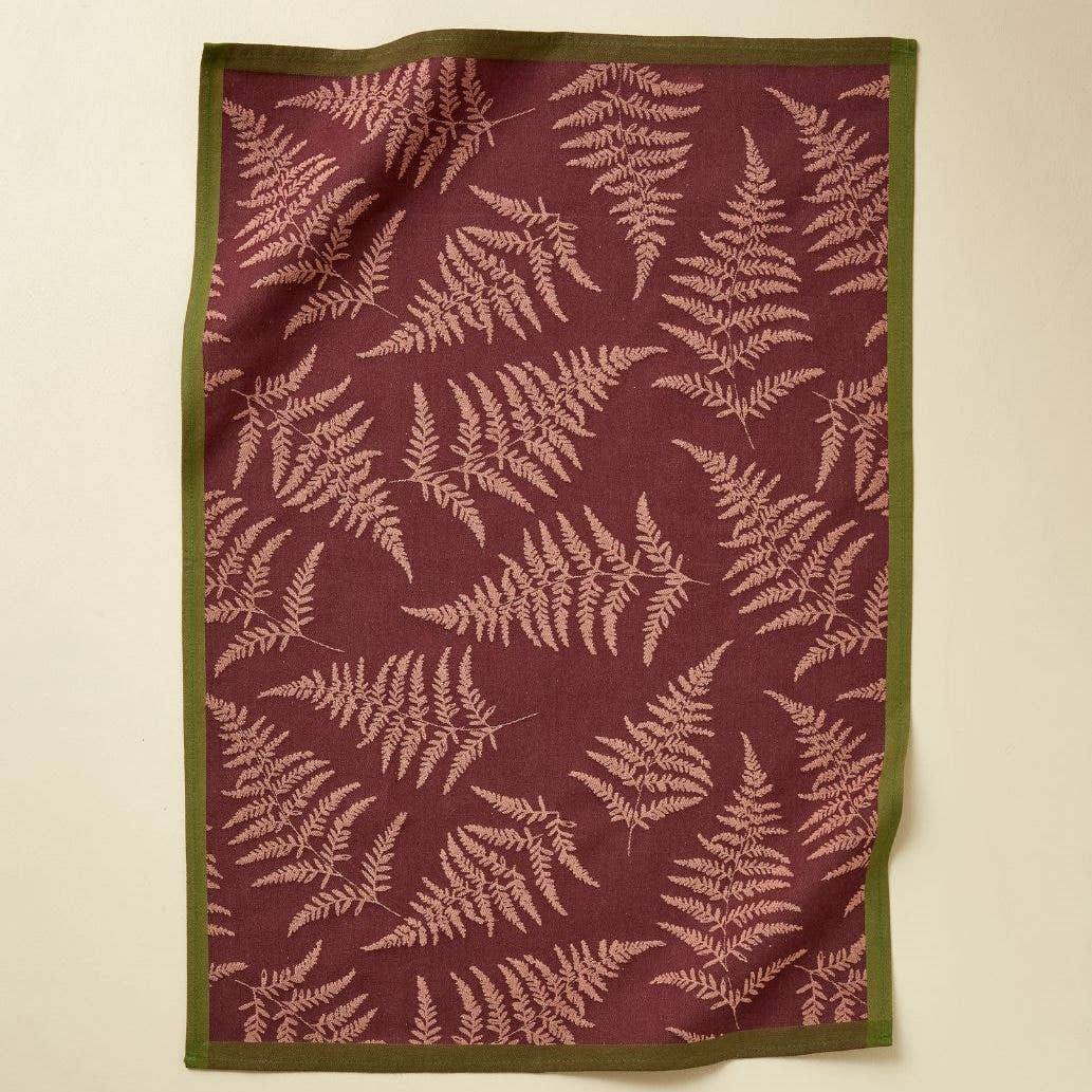 Ein Sylvie Thiriez - Geschirrtücher Jacquard Verzauberter Wald Bordeaux Geschirrtuch mit kastanienbraunem Hintergrund, hellrosa Farnmotiv und olivgrüner Bordüre liegt flach auf einer hellen Küchenfläche.