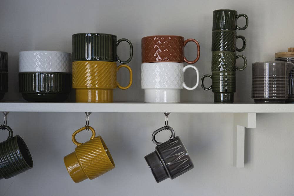 Auf einem weißen Wandregal sind gestapelte Kaffeebecher (2-teilig) der Sagaform GmbH Coffee & More in Grün, Weiß, Senfgelb, Braun und Schwarz ausgestellt. Darunter hängen drei Tassen mit geometrischen Mustern an ihren Henkeln.