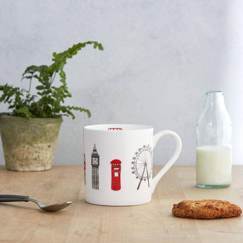 Der Victoria Eggs - London Skyline Becher von Victoria Eggs, ein Becher aus feinem Knochenporzellan mit Londoner Wahrzeichen, steht mit einem Keks, einer Milchflasche, einem Löffel und einer Topfpflanze auf Holz – ein bezauberndes britisches Souvenir für jeden Fan.