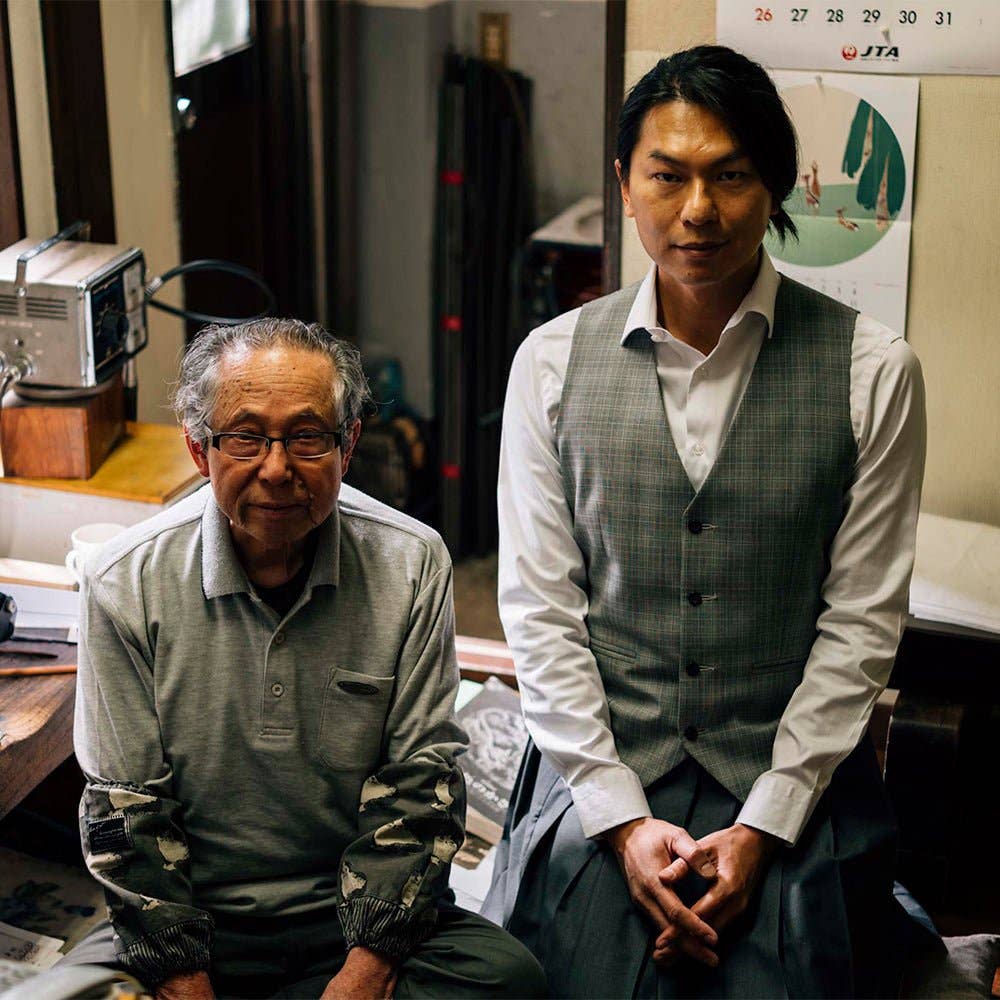 Ein älterer Mann mit Brille und grauem Hemd sitzt neben einem jüngeren Mann mit weißem Hemd und grauer Weste in einem Haus. Auf dem Tisch liegen ein Kalender, ein Radio und ein ENTREPÔT - YSTUDIO Klassische Renaissance KAZARI KANAGU ORCHID Tintenroller-Stift.