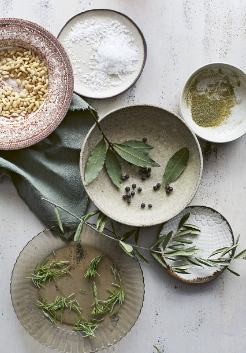 Fünf Keramikschalen auf einer hellen Oberfläche enthalten Pinienkerne, Salz, Kräuter, Pfefferkörner und Rosmarinzweige - perfekt für Rezepte aus "How to eat your christmas tree" vom Ars Vivendi Verlag. Unter den Schalen ist teilweise ein grünes Tuch zu sehen.