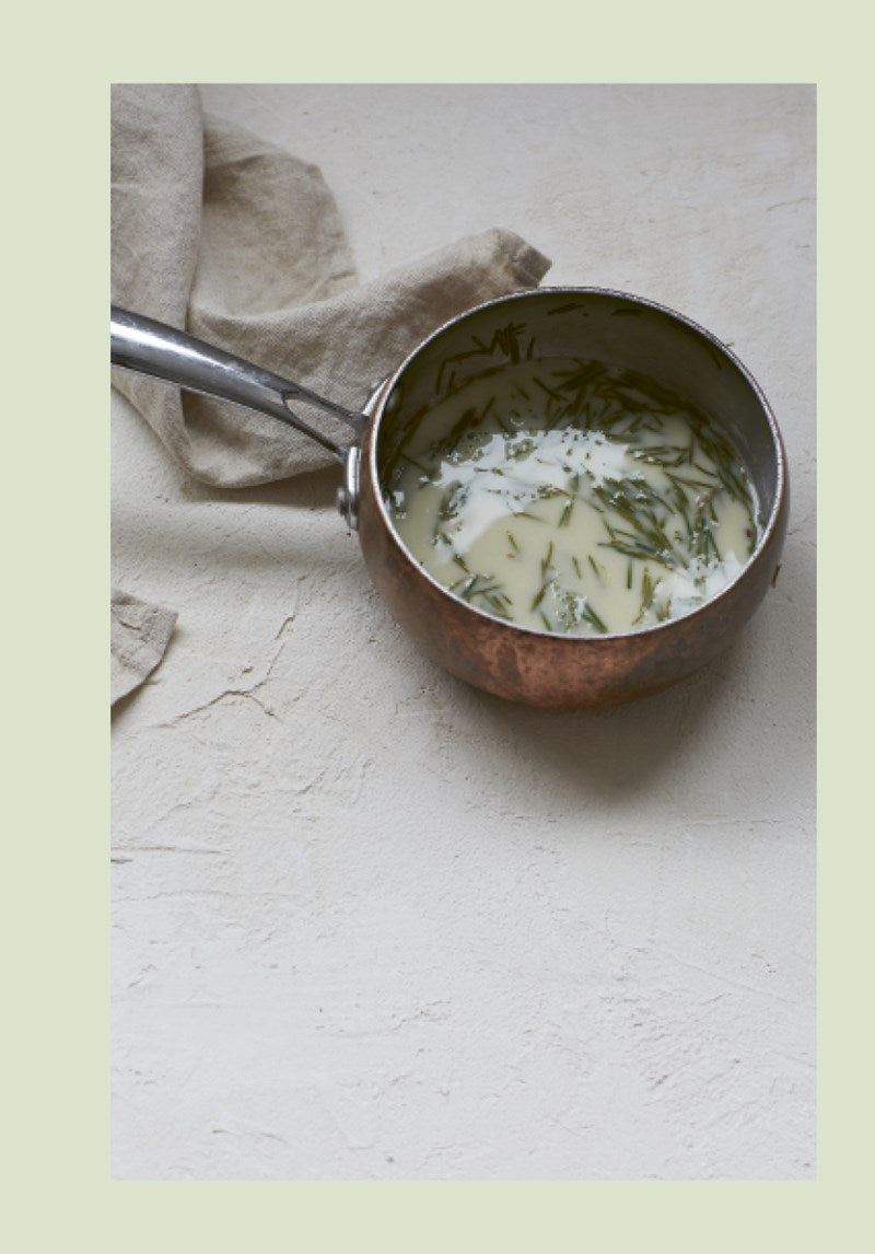 Ein Kupfertopf mit cremiger Flüssigkeit und frischen Kräutern - perfekt für Rezepte aus "How to eat your christmas tree" vom Ars Vivendi Verlag - steht auf einer hellen Fläche neben einer beigen Serviette und erinnert an ein nachhaltiges Kochbuch.