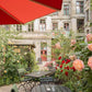 Genießen Sie eine gemütliche Gartencafé-Terrasse, wie sie in "Stadtoasen in Berlin" vom Bebra Verlag abgebildet ist, mit schwarzen Tischen und Stühlen inmitten von blühenden Blumen unter einem roten Sonnenschirm, vor einem alten Gebäude mit rot gerahmten Fenstern.