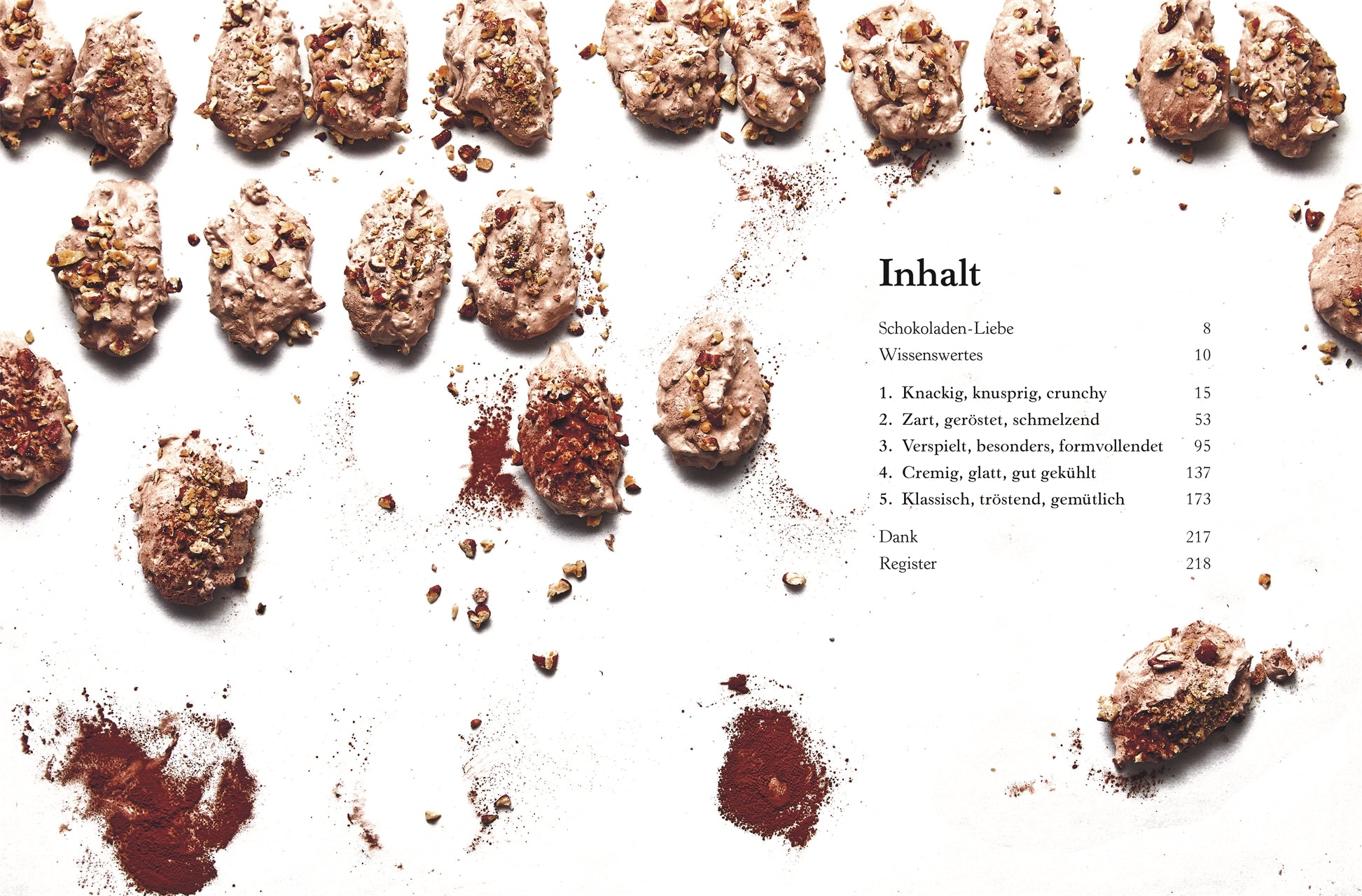Reihen von schokoladenüberzogenen Nuss-Clustern auf weißem, mit Kakaopulver bestäubtem Hintergrund. Rechts ist ein deutsches Inhaltsverzeichnis von „Alles aus Schokolade“ des DK Verlags mit Rezepten und Seitenzahlen zu sehen, darunter sind Kakaopulverflecken zu sehen.