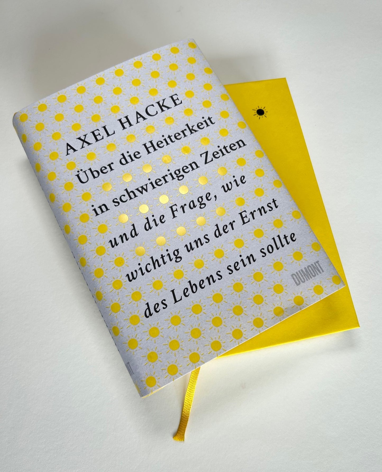 Das von Friedrich Schiller inspirierte Buch Über die Heiterkeit aus dem Dumont Verlag liegt teilweise aufgeschlagen auf einer weißen Fläche mit gelb-grauem, sonnengemustertem Einband.