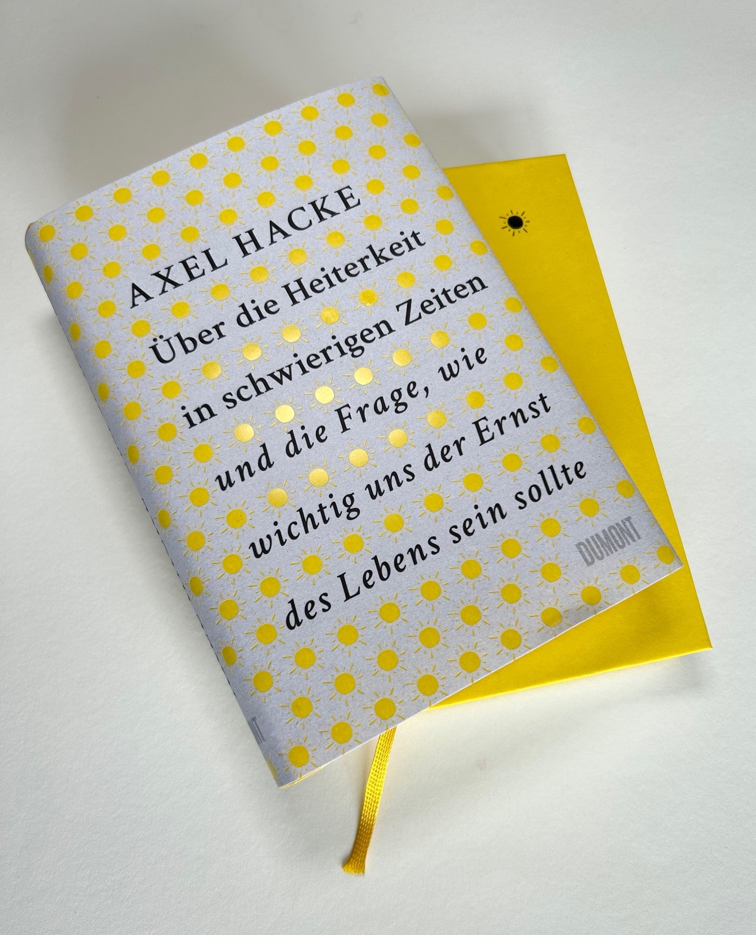 Das von Friedrich Schiller inspirierte Buch Über die Heiterkeit aus dem Dumont Verlag liegt teilweise aufgeschlagen auf einer weißen Fläche mit gelb-grauem, sonnengemustertem Einband.